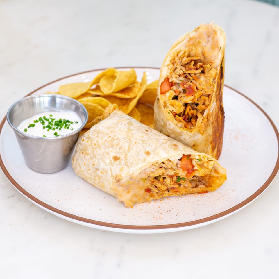 Two grilled chicken burrito halves with rice and vegetables, tortilla chips, and sour cream topped with chives on a plate.
