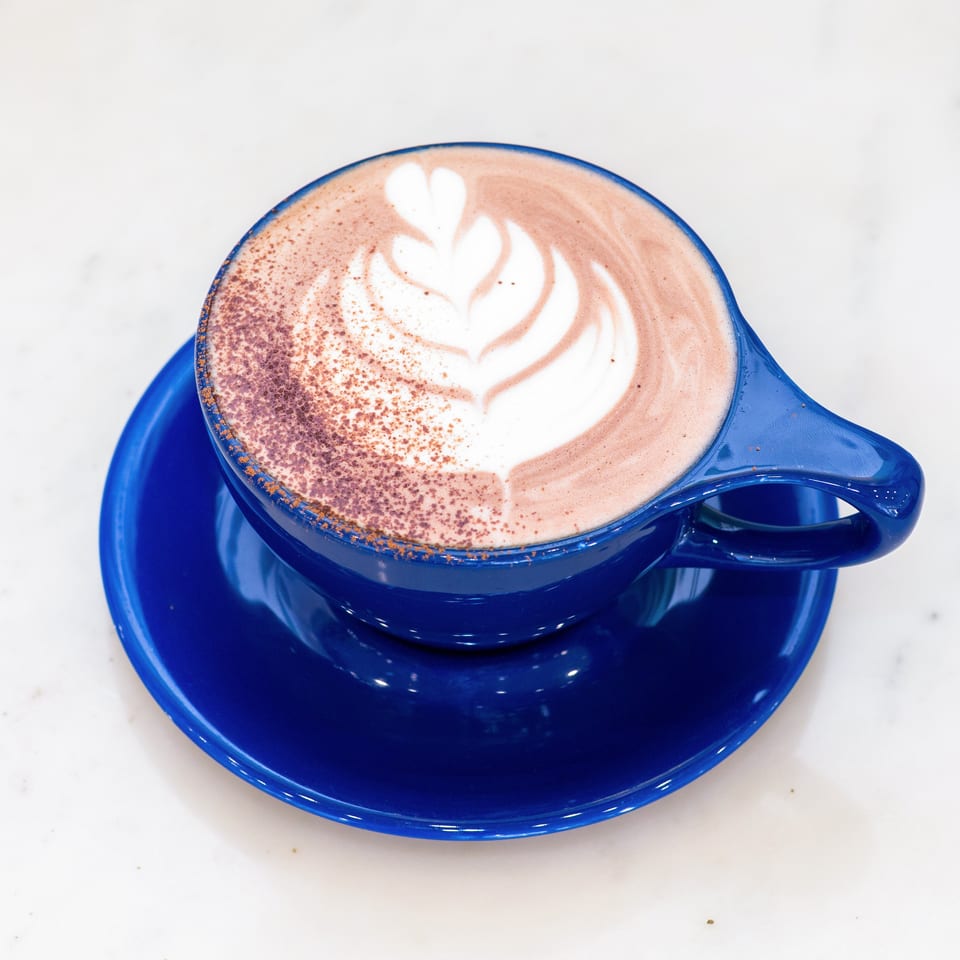 Cocoa-dusted latte with heart-shaped foam art in a bright blue cup and saucer.