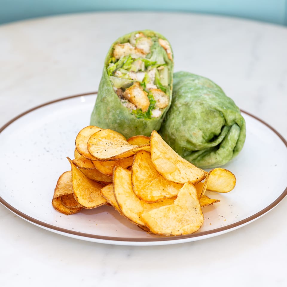 Green wrap filled with lettuce, chicken, cucumber, and croutons served with golden potato chips on a white plate.