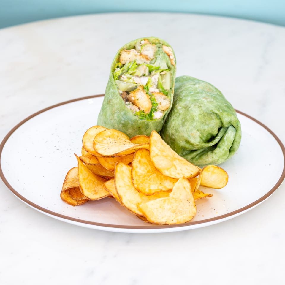 Green wrap filled with lettuce, chicken, and croutons served with golden potato chips on a white plate.