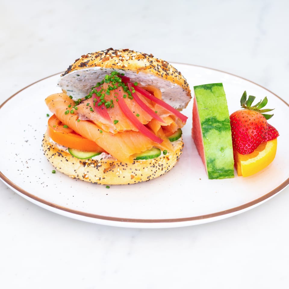 Everything bagel sandwich layered with smoked salmon, cream cheese, tomatoes, cucumber, onions, and chives, served with watermelon and strawberries.