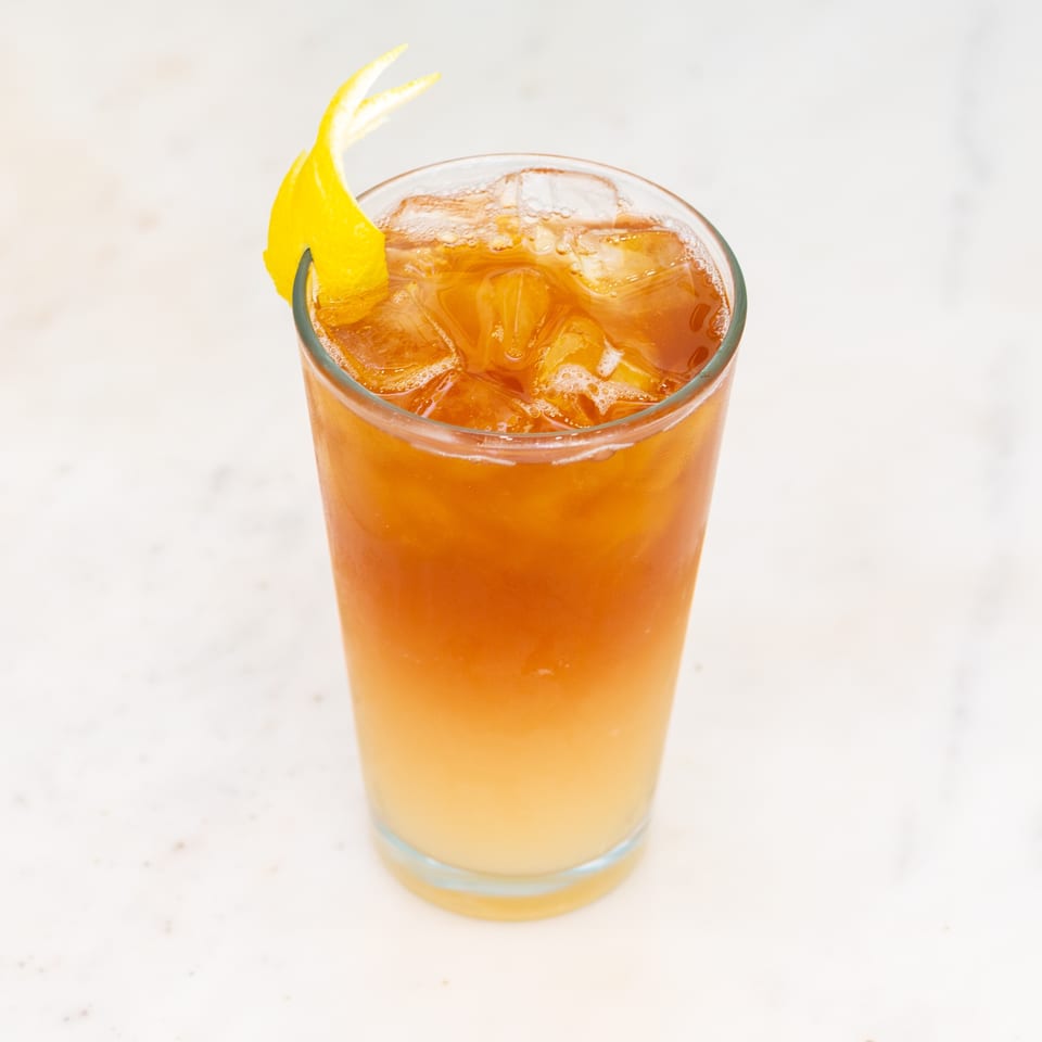 Amber iced cocktail in a clear glass, garnished with a twisted lemon peel on a light countertop.