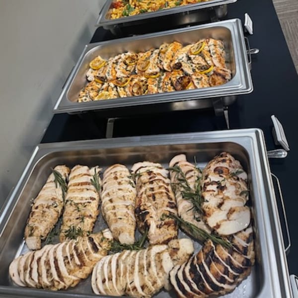Three chafing dishes filled with grilled chicken, one with lemon slices, and another with rosemary.