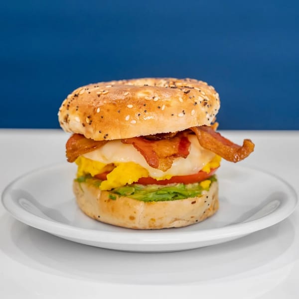 Everything bagel sandwich with bacon, egg, cheese, tomato, and avocado on a white plate.