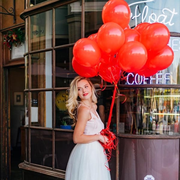 Blonde woman in a white dress holding red balloons outside Float Restaurant, with a neon sign in the window.