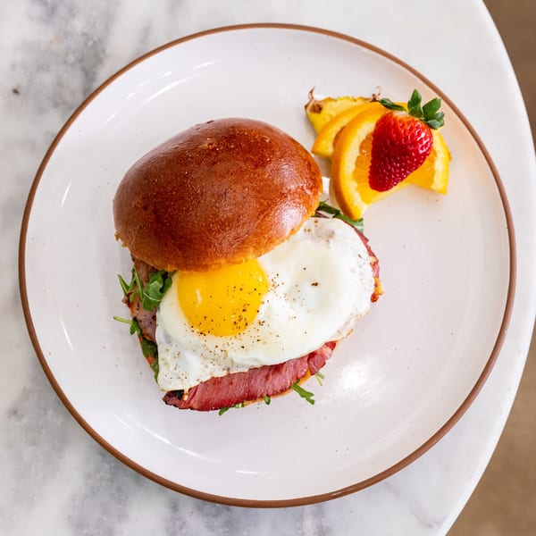 A breakfast sandwich with a fried egg, meat, and arugula, served with fruit on a white plate.