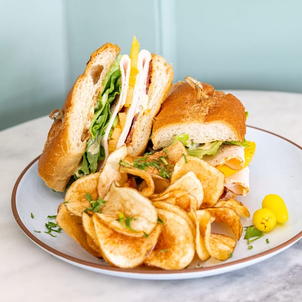 Float Restaurant's turkey sandwich on a baguette with lettuce, cheese, and chips on a white plate.