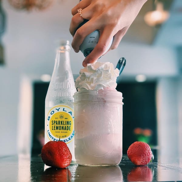 A hand adds whipped cream to a Float Restaurant strawberry float next to a bottle of sparkling lemonade.