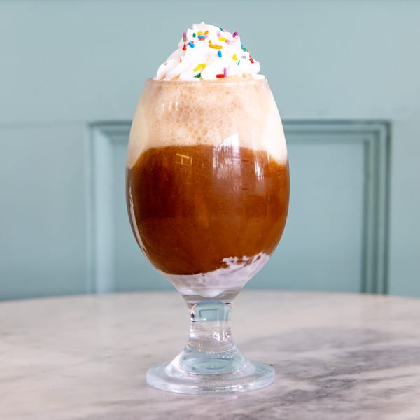 A root beer float with whipped cream and colorful sprinkles sits on a marble table.